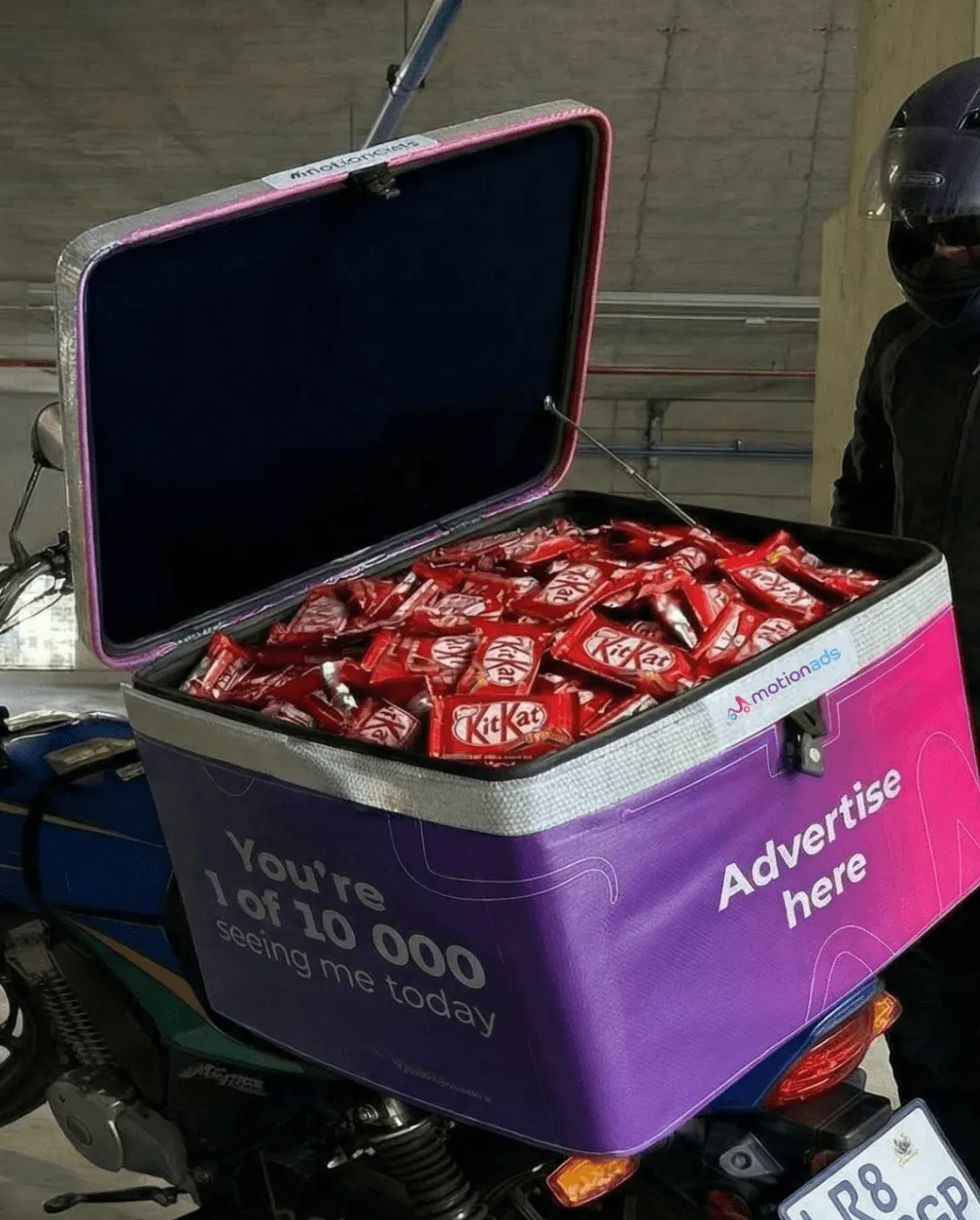 An open MotionAds top-box loaded with stock on the back of a delivery motorbike, its purple and pink wrap reading Advertise here.